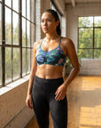A woman standing in a bright, sunlit studio with large windows, wearing a ruched sports bra with a teal, green, and purple abstract print, paired with black leggings.
